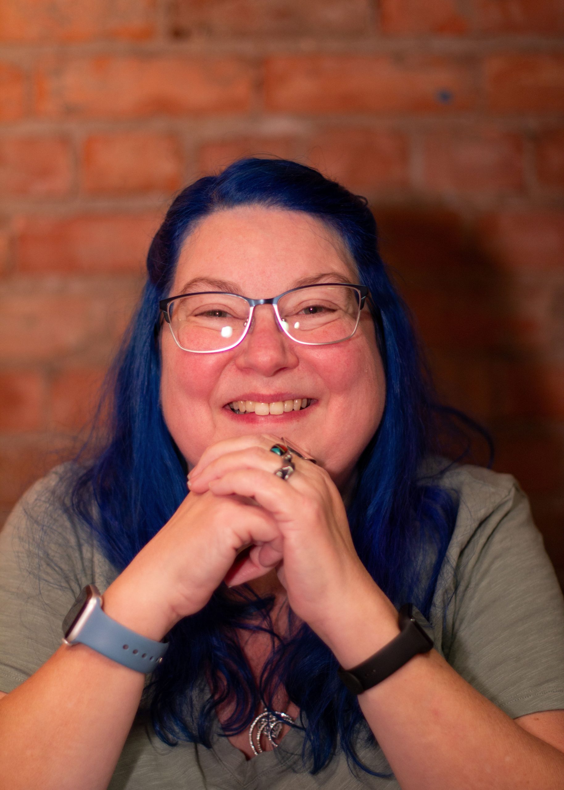 Picture of a woman with blue hair, smiling, with her chin behind her linked hands