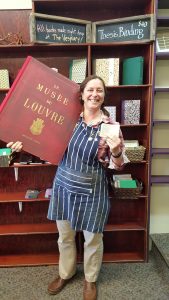 Audra holding a huge art book and a tiny book in front of shelves holding handmade journals